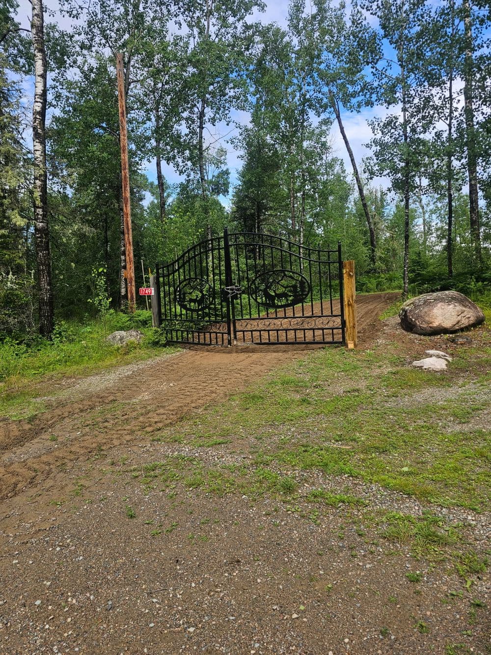 Black wrought-iron gate on a gravel path surrounded by lush green trees and rocks.