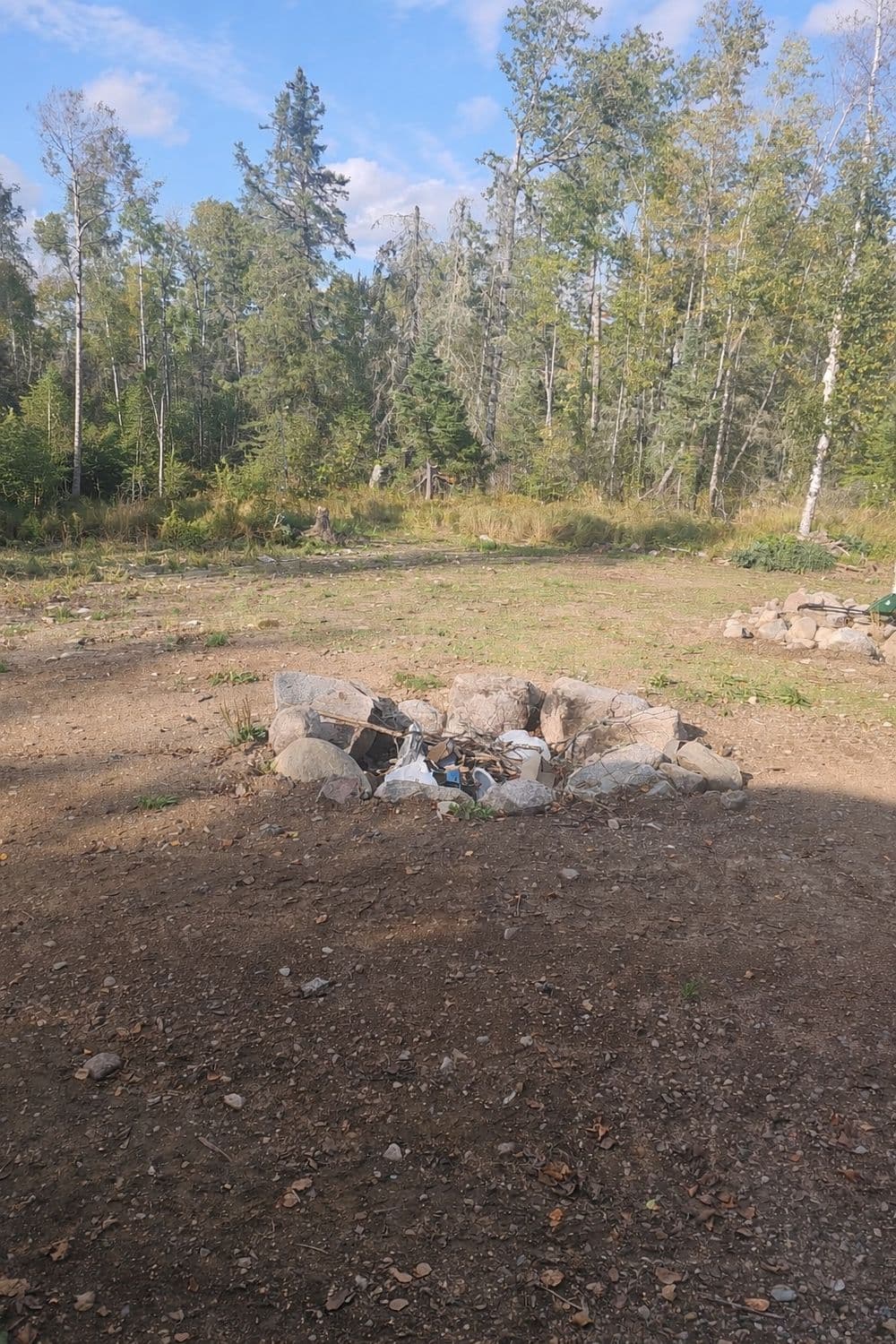 Outdoor campsite featuring a stony fire pit surrounded by forested area and clear blue sky.
